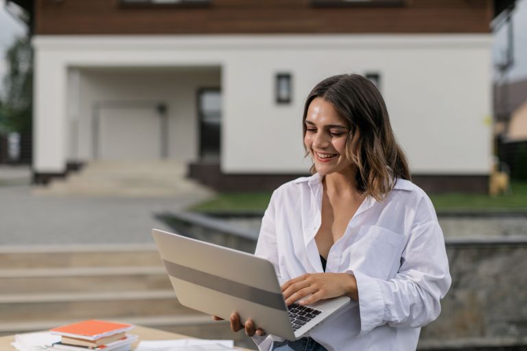 Careers In Real Estate professional smiling while working on a laptop in front of a house for sale, representing career opportunities and growth within Australia's real estate industry.