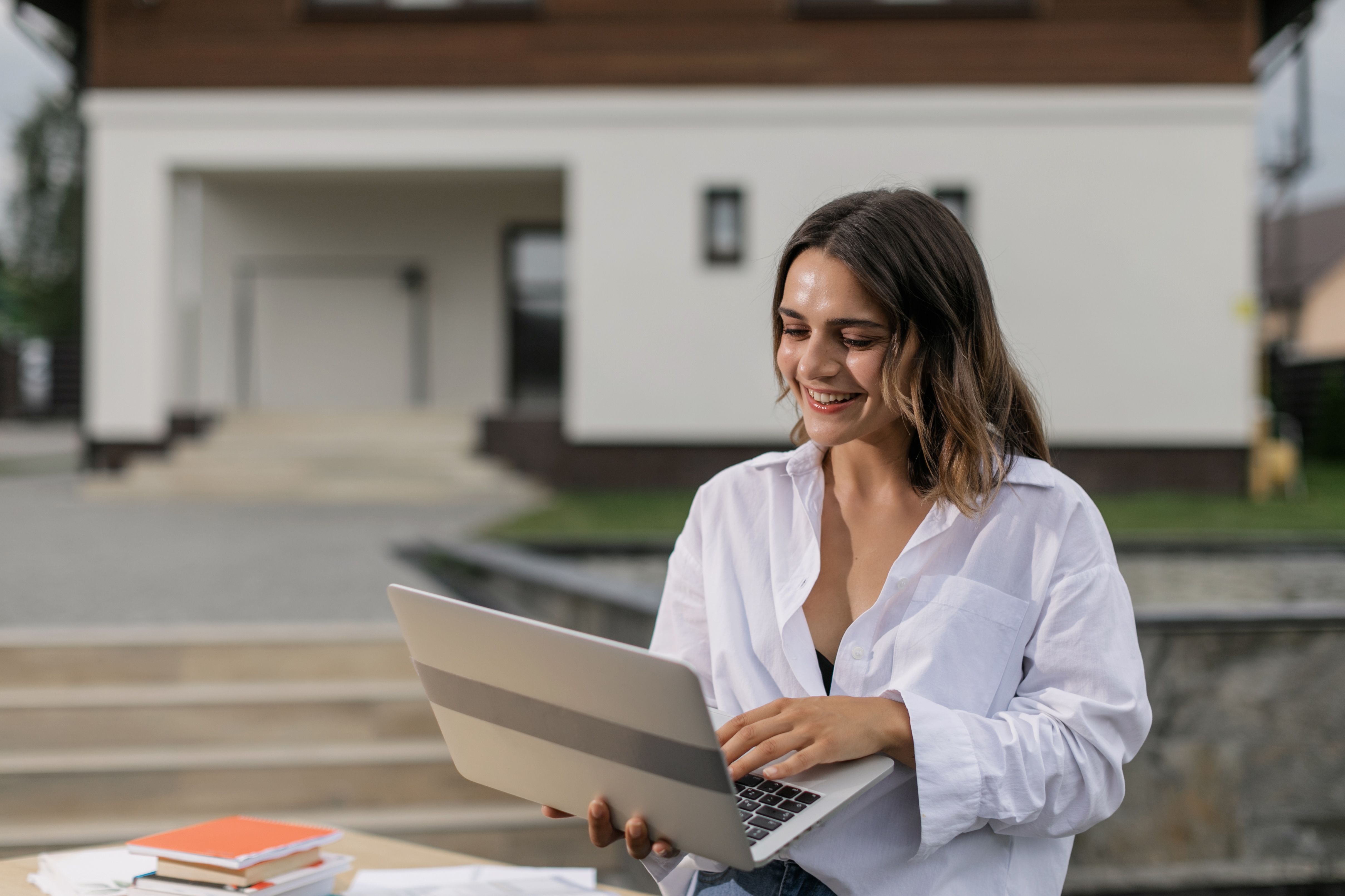 Careers In Real Estate professional smiling while working on a laptop in front of a house for sale, representing career opportunities and growth within Australia's real estate industry.